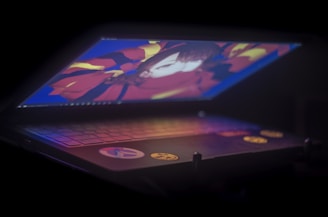 A dimly lit laptop partially opened, displaying colorful anime-style artwork on its screen. The keyboard is visible with some decorative stickers.
