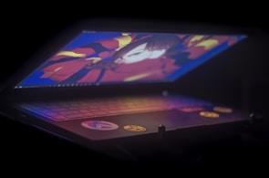 A dimly lit laptop partially opened, displaying colorful anime-style artwork on its screen. The keyboard is visible with some decorative stickers.