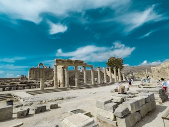 Ancient ruins with large stone columns and scattered stones can be seen. A few tourists are present, some walking and others sitting, all exploring the site. A tree stands near the center of the ruins. The sky is clear with some clouds, suggesting a sunny day.
