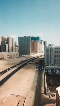 A cityscape featuring a wide highway with minimal traffic running alongside a construction site. Tall buildings with modern architecture dominate the skyline under a clear blue sky. The foreground shows an almost barren sandy area with a few construction barriers.