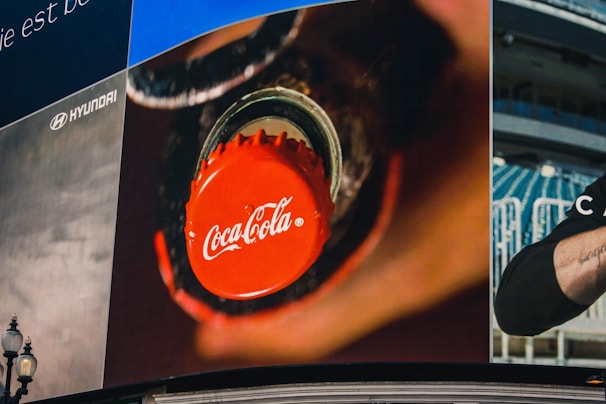 A large billboard displays a close-up of a Coca-Cola bottle cap being unscrewed. The cap is bright red with the iconic Coca-Cola logo in white. In the background, there are hints of a stadium setting with seats visible, as well as a partial view of a person's arm.