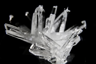 A cluster of transparent, angular crystal formations against a dark background. The edges of the crystals catch the light, creating a gleaming effect. The irregular arrangement of the crystals gives a sense of natural beauty and complexity.