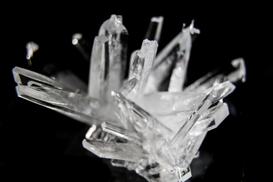 A cluster of transparent, angular crystal formations against a dark background. The edges of the crystals catch the light, creating a gleaming effect. The irregular arrangement of the crystals gives a sense of natural beauty and complexity.