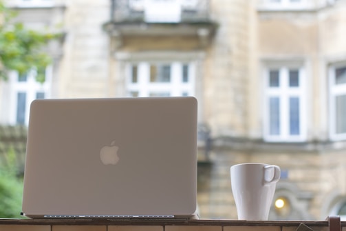 A laptop with a logo on its lid sits on a ledge beside a white coffee mug. Visible in the background is a building with beige stone walls and large white-framed windows. The scene appears to be taken outdoors or near a window, with soft natural lighting.