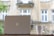 A laptop with a logo on its lid sits on a ledge beside a white coffee mug. Visible in the background is a building with beige stone walls and large white-framed windows. The scene appears to be taken outdoors or near a window, with soft natural lighting.