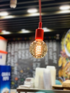 A glowing light bulb with a red fixture is prominently hanging in focus, with a blurred background featuring a chalkboard covered in text and colorful imagery. The scene has a cozy, warm ambiance reminiscent of a cafe or casual dining area.