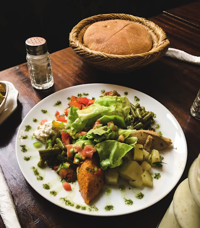 A plate of vibrant, colorful food featuring a variety of vegetables, including lettuce, cucumber, green beans, and diced tomatoes, is presented. There is a mix of cooked and fresh items, such as slices of a patty or loaf and small potato cubes, garnished with herbs or spices. A wicker basket containing a whole round bread sits on the table beside a salt shaker.