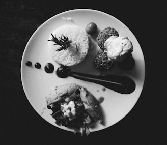 A monochrome image featuring a neatly arranged gourmet meal on a round white plate. The composition includes a portion of rice topped with a sprig of rosemary, accompanied by falafel balls topped with sauce, a piece of pita filled with vegetables, a small tomato, and artistic splatters of sauce.