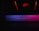A glowing, backlit logo with an abstract design in red is positioned on the top portion of the image. Below, a dimly lit surface displays a soft gradient of purple, blue, and pink hues, suggesting an illuminated keyboard or similar device.