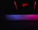 A glowing, backlit logo with an abstract design in red is positioned on the top portion of the image. Below, a dimly lit surface displays a soft gradient of purple, blue, and pink hues, suggesting an illuminated keyboard or similar device.