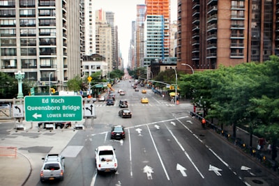 A bustling urban street with tall buildings on either side, featuring numerous vehicles, including yellow taxis and a road sign indicating directions to the Ed Koch Queensboro Bridge. The street is lined with green trees and pedestrians are visible along the sidewalks.