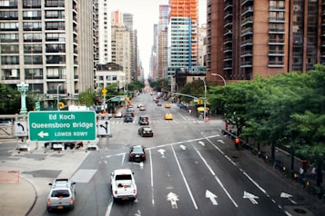 A bustling urban street with tall buildings on either side, featuring numerous vehicles, including yellow taxis and a road sign indicating directions to the Ed Koch Queensboro Bridge. The street is lined with green trees and pedestrians are visible along the sidewalks.