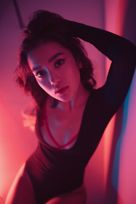 A person poses with one arm raised, wearing a black outfit with visible red straps. The image is illuminated with dramatic red and blue lighting, creating a moody and artistic effect. The background is a plain wall, and the person gazes directly at the camera with a serious expression.