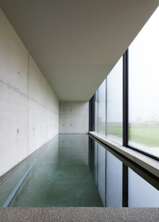 A modern and minimalist interior space featuring a long rectangular pool of water alongside a large window. The design is characterized by clean lines, polished concrete surfaces, and an overall sense of tranquility. Natural light softly illuminates the room through the window, creating reflections on the water.