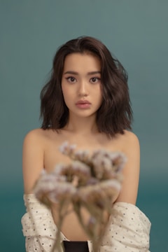 A young woman with wavy dark hair gazes directly at the camera against a teal background. She is wearing an off-the-shoulder polka dot robe, and a soft-focus bouquet of flowers is positioned in the foreground.