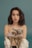 A young woman with wavy dark hair gazes directly at the camera against a teal background. She is wearing an off-the-shoulder polka dot robe, and a soft-focus bouquet of flowers is positioned in the foreground.