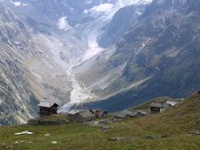 A picturesque mountain valley with snow-capped peaks and a winding river carving through the landscape. In the foreground, there are several rustic wooden cabins on a grassy slope.