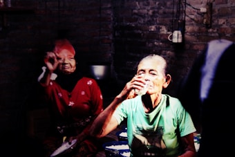 An elderly woman wearing a light green shirt drinks from a glass, while another elderly woman in a red dress sits behind her in a dimly lit room with exposed brick walls.