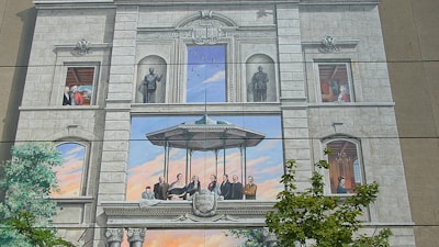 The image displays a detailed mural that looks like an architectural facade with realistic painted windows and balconies. It features a Renaissance-style structure with people depicted in various historical or formal attire. Statues and columns add to the grandeur, and a central scene includes individuals gathered under a pavilion. There are elements of nature, such as a tree, blending with the architectural painting.