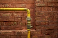 A yellow metal pipe runs horizontally along a red brick wall with a 90-degree elbow joint, showing signs of age and wear.