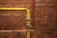 A yellow metal pipe runs horizontally along a red brick wall with a 90-degree elbow joint, showing signs of age and wear.