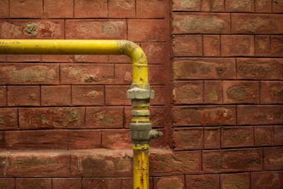 A yellow metal pipe runs horizontally along a red brick wall with a 90-degree elbow joint, showing signs of age and wear.