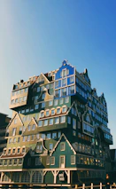 A uniquely designed building composed of stacked, colorful modular units resembling traditional Dutch houses. The architecture features multiple roof styles and windows, adding to the eclectic appearance. The sky is clear with a warm, soft light illuminating the structure.
