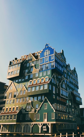 A uniquely designed building composed of stacked, colorful modular units resembling traditional Dutch houses. The architecture features multiple roof styles and windows, adding to the eclectic appearance. The sky is clear with a warm, soft light illuminating the structure.