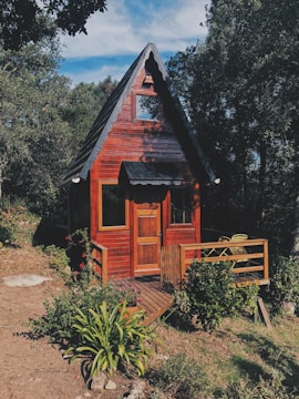 A charming wooden cabin with an A-frame roof is nestled among lush green trees. The cabin features a small porch with a couple of chairs, surrounded by vibrant bushes and plants.