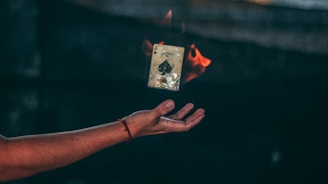 A hand is extended in mid-air, palm facing up, with a burning playing card, specifically the ace of spades, floating above it. The card is engulfed in flames on one side, creating a dramatic effect against the dark, blurred background.