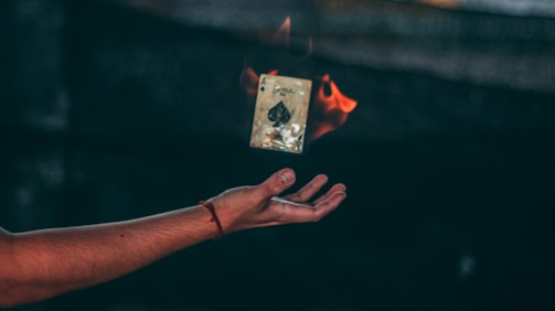 A hand is extended in mid-air, palm facing up, with a burning playing card, specifically the ace of spades, floating above it. The card is engulfed in flames on one side, creating a dramatic effect against the dark, blurred background.