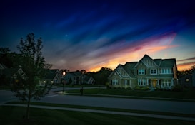 A suburban neighborhood at dusk, featuring a large, well-lit house with multiple windows and a landscaped front yard. The sky is a blend of dark blue and soft orange hues, suggesting a sunset. A streetlamp is illuminated, casting a warm glow on the pavement, and a tree is visible on the left side of the image.