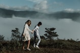 A couple walks hand in hand along a grassy hilltop with foggy mountains in the background. The woman wears a floral dress and holds a bouquet of flowers, while the man is dressed in casual attire. The scene is serene and romantic, with trees and lush greenery surrounding them.