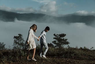 A couple walks hand in hand along a grassy hilltop with foggy mountains in the background. The woman wears a floral dress and holds a bouquet of flowers, while the man is dressed in casual attire. The scene is serene and romantic, with trees and lush greenery surrounding them.