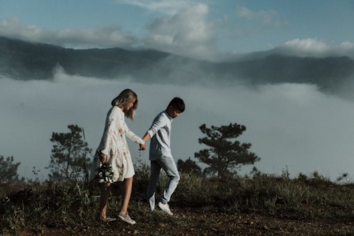 A couple walks hand in hand along a grassy hilltop with foggy mountains in the background. The woman wears a floral dress and holds a bouquet of flowers, while the man is dressed in casual attire. The scene is serene and romantic, with trees and lush greenery surrounding them.