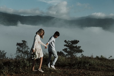 A couple walks hand in hand along a grassy hilltop with foggy mountains in the background. The woman wears a floral dress and holds a bouquet of flowers, while the man is dressed in casual attire. The scene is serene and romantic, with trees and lush greenery surrounding them.
