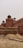 A historic fort made of red sandstone, featuring large walls, domed structures, and a series of arches. The architecture reflects ancient Indian designs with symmetrical patterns and intricate details. In the foreground, there are piles of sand and barriers under a cloudy sky, suggesting ongoing construction or restoration work.
