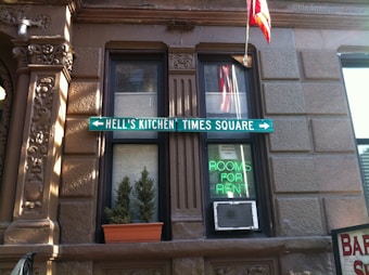 A brick building facade with two windows features a green directional sign pointing to Hell's Kitchen and Times Square. One window has a small potted plant, and the other displays a neon sign advertising 'Rooms for Rent' above an air conditioning unit. An American flag is visible above.