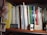 A bookshelf filled with a variety of books, magazines, and folders. To the left, there is a yellow folder labeled 'English.' A stack of magazines is placed vertically, alongside various books with colorful spines. Some titles include 'International & Foreign Programs' and 'Guide to Proposal Writing.' A small 'New York Wendy' license plate is displayed on the shelf, accompanied by a model car, a pack of cards, and a NYC event badge. A small figurine or decorative item is on the far right.