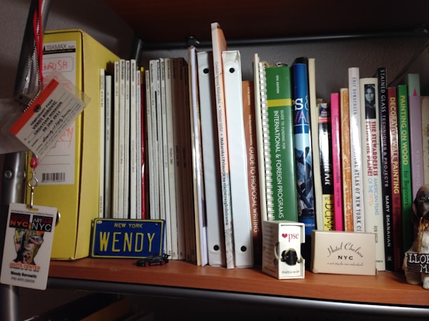 A bookshelf filled with a variety of books, magazines, and folders. To the left, there is a yellow folder labeled 'English.' A stack of magazines is placed vertically, alongside various books with colorful spines. Some titles include 'International & Foreign Programs' and 'Guide to Proposal Writing.' A small 'New York Wendy' license plate is displayed on the shelf, accompanied by a model car, a pack of cards, and a NYC event badge. A small figurine or decorative item is on the far right.