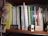A bookshelf filled with a variety of books, magazines, and folders. To the left, there is a yellow folder labeled 'English.' A stack of magazines is placed vertically, alongside various books with colorful spines. Some titles include 'International & Foreign Programs' and 'Guide to Proposal Writing.' A small 'New York Wendy' license plate is displayed on the shelf, accompanied by a model car, a pack of cards, and a NYC event badge. A small figurine or decorative item is on the far right.
