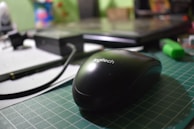 A close-up of a black Logitech computer mouse on a green cutting mat. The background includes a laptop and various office supplies, such as a pen and a highlighter, which are slightly out of focus.
