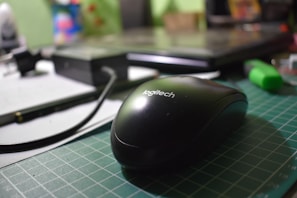 A close-up of a black Logitech computer mouse on a green cutting mat. The background includes a laptop and various office supplies, such as a pen and a highlighter, which are slightly out of focus.