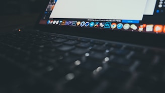 A close-up view of an open laptop displaying a variety of application icons on the desktop. The screen is partially visible with a document open, while the keyboard is softly lit and slightly out of focus, creating a dim and moody atmosphere.