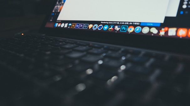 A close-up view of an open laptop displaying a variety of application icons on the desktop. The screen is partially visible with a document open, while the keyboard is softly lit and slightly out of focus, creating a dim and moody atmosphere.