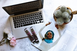 A flat lay composition featuring an open laptop, a magazine with a page showing a woman in vibrant attire, two chocolate bars, a potted cactus, and a small bouquet of roses placed on a textured white surface.