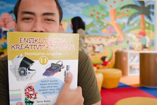 A person holds up a children's encyclopedia titled 'Ensiklopedia Kreativitas Anak' in front of their face. The cover features images related to space exploration, such as a helmet and spacecraft tools. The background displays a colorful mural of a playful outdoor scene with trees, clouds, and abstract figures.