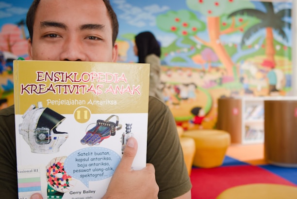 A person holds up a children's encyclopedia titled 'Ensiklopedia Kreativitas Anak' in front of their face. The cover features images related to space exploration, such as a helmet and spacecraft tools. The background displays a colorful mural of a playful outdoor scene with trees, clouds, and abstract figures.