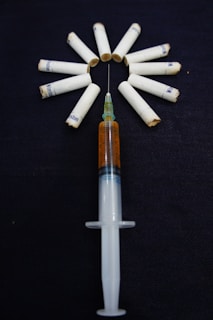 A syringe filled with a brown liquid is placed on a dark surface, surrounded by several cigarette filters arranged in a circle. The arrangement seems intentional and creates a striking visual with the cigarettes fanning out from the needle.