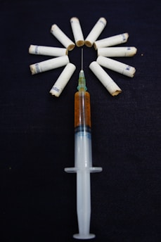 A syringe filled with a brown liquid is placed on a dark surface, surrounded by several cigarette filters arranged in a circle. The arrangement seems intentional and creates a striking visual with the cigarettes fanning out from the needle.